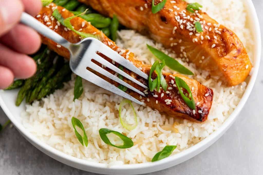 A person holding a fork in a teriyaki salmon strips with rice and green toppings on a plate.