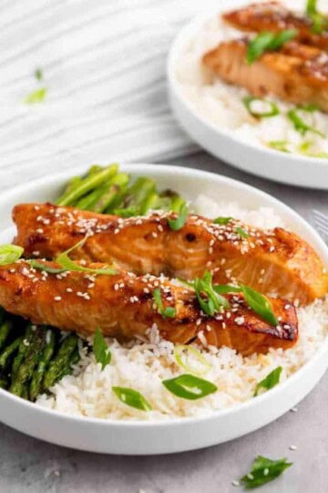 Two teriyaki salmon strips with rice and green toppings on a plate.