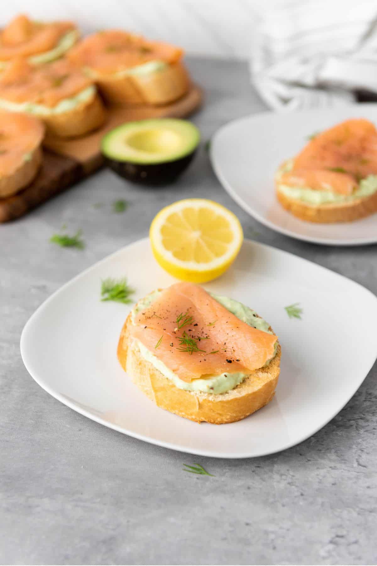 A piece of smoked salmon and avocado bruschetta with sliced lemon nearby.