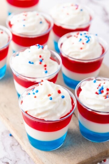 A tray of jell-O shots in small glasses showing blue, white, and red layers, topped with whipped cream and sprinkles.