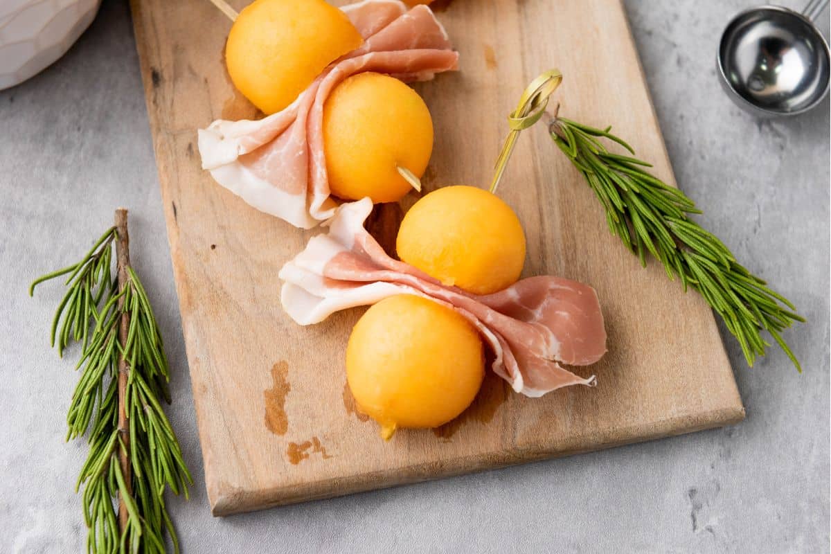 Cantaloupe Prosciutto Skewers on a wooden board with rosemary sprigs.