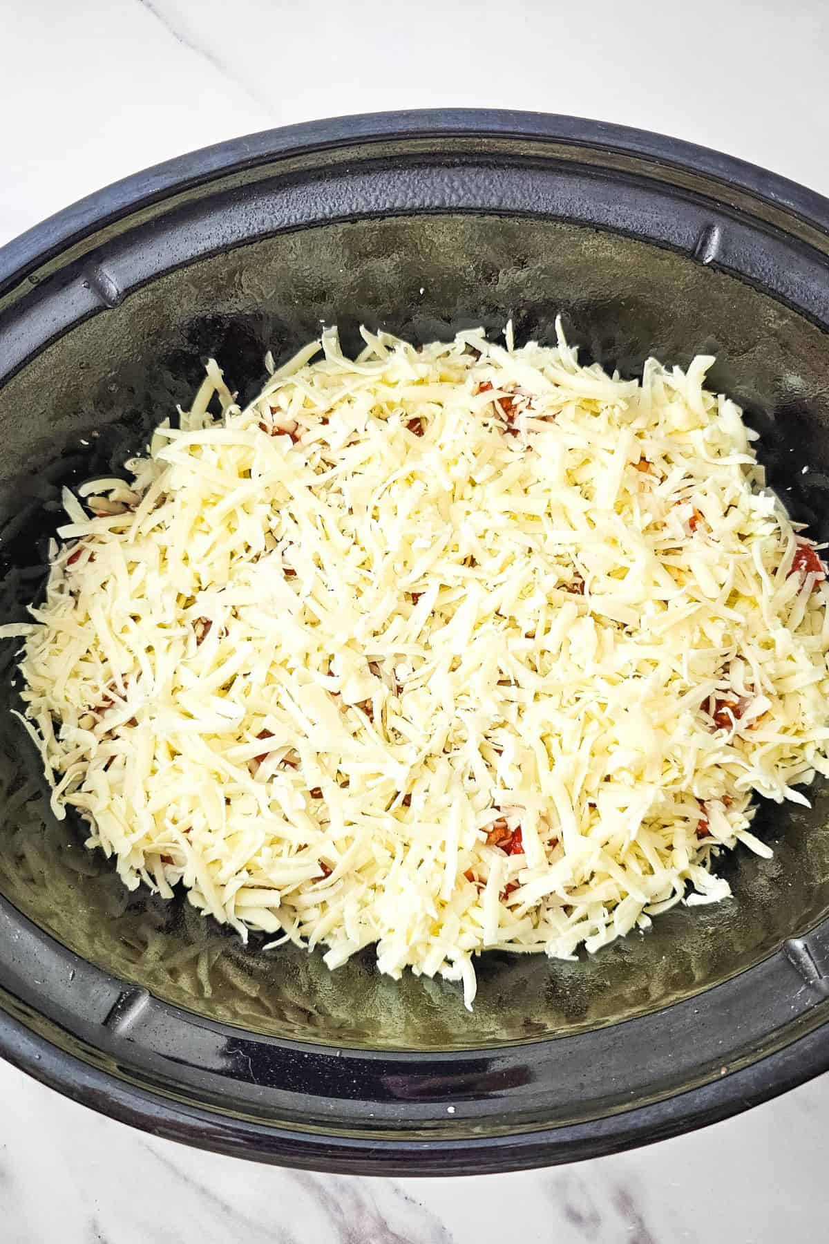 Lasagna in the crockpot being topped with shredded cheese, ready to cook.