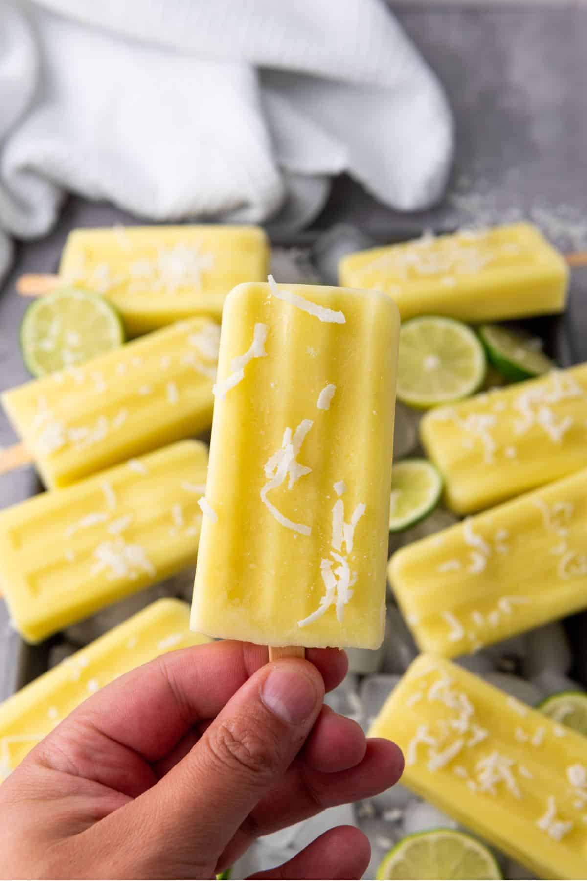 A hand holding a single pineapple coconut lime popsicle with more popsicles in the background.