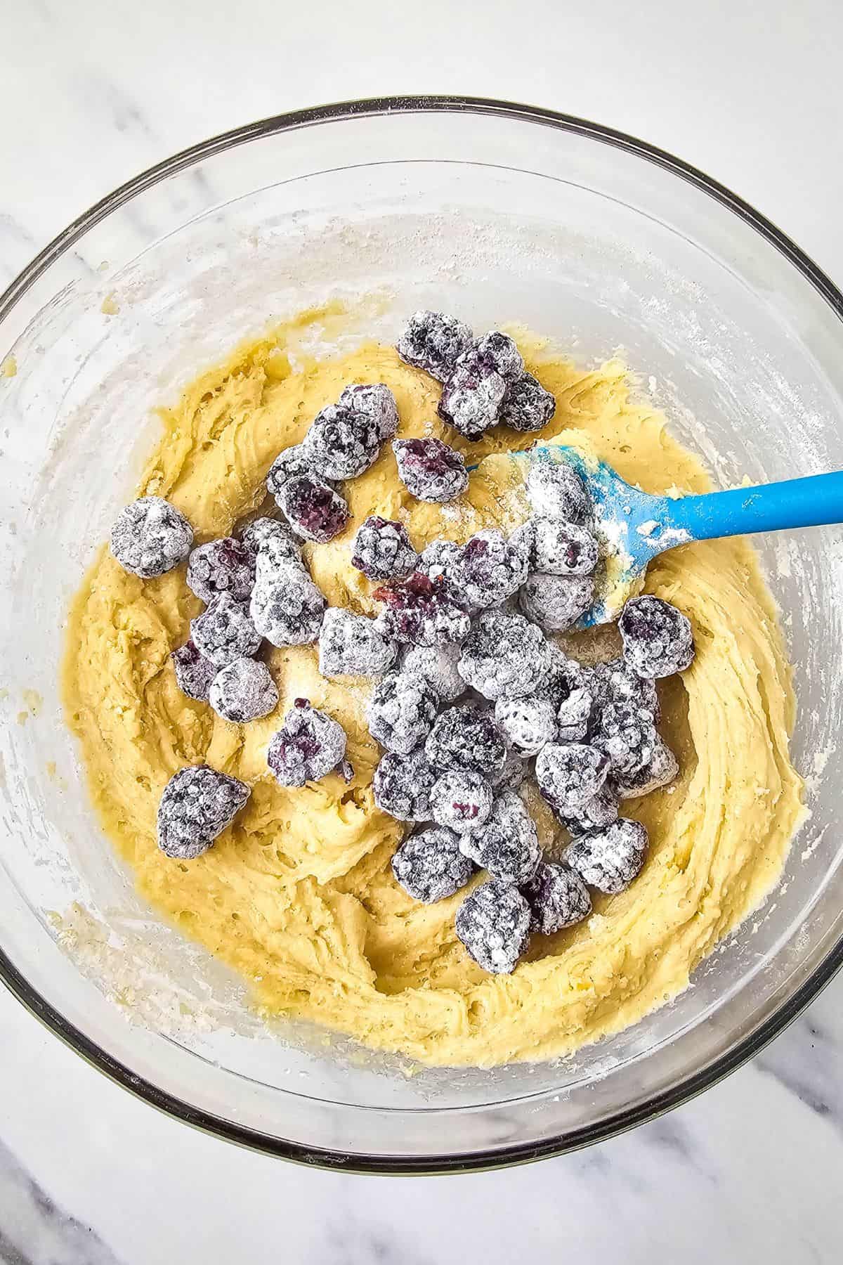 A mixing bowl filled with batter, topped with flour-coated blueberries and a spatula resting inside.