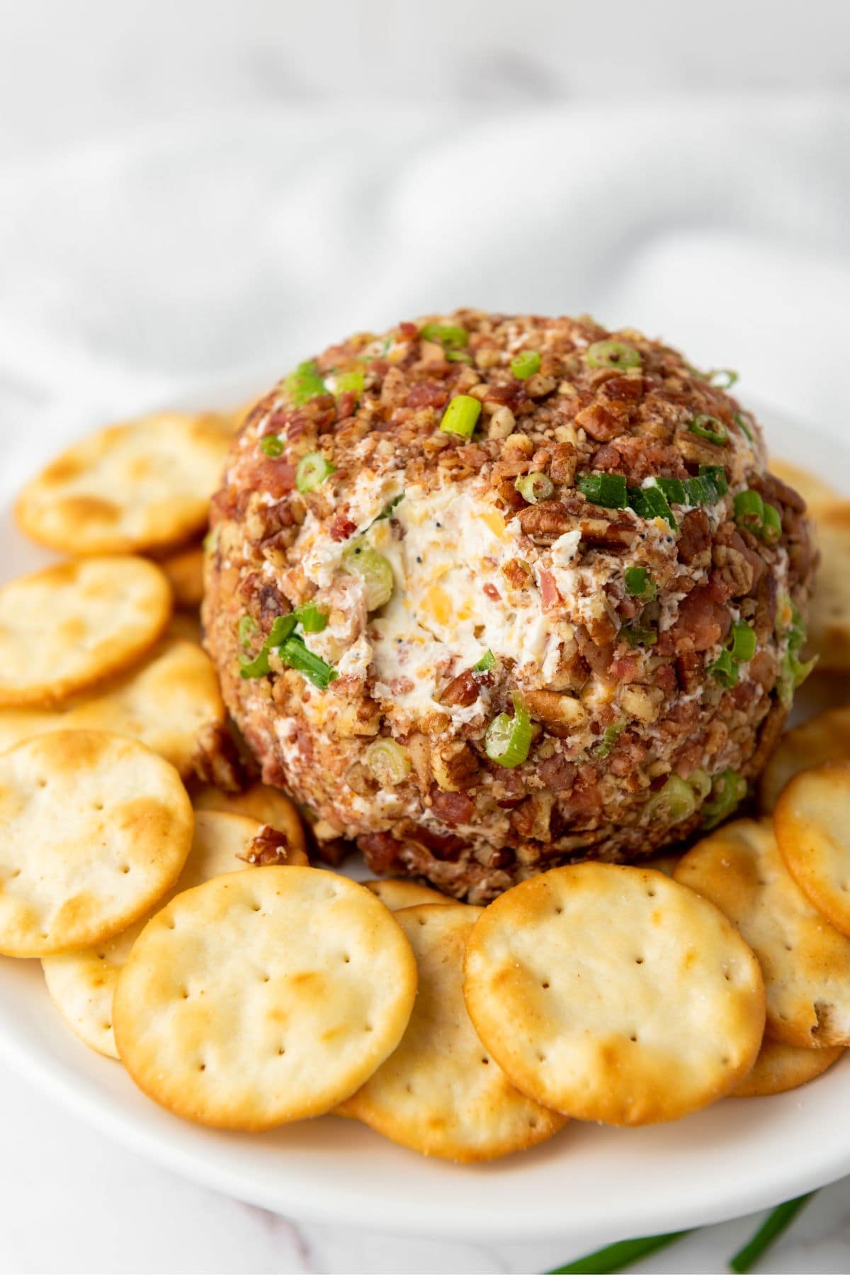 A round cheeseball coated with chopped pecans, bacon bits, and scallions, placed on a plate and surrounded by crackers.