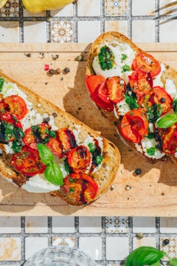 Two pieces of burrata toast on a plate, topped with roasted tomatoes, fresh basil, and a drizzle of basil sauce.