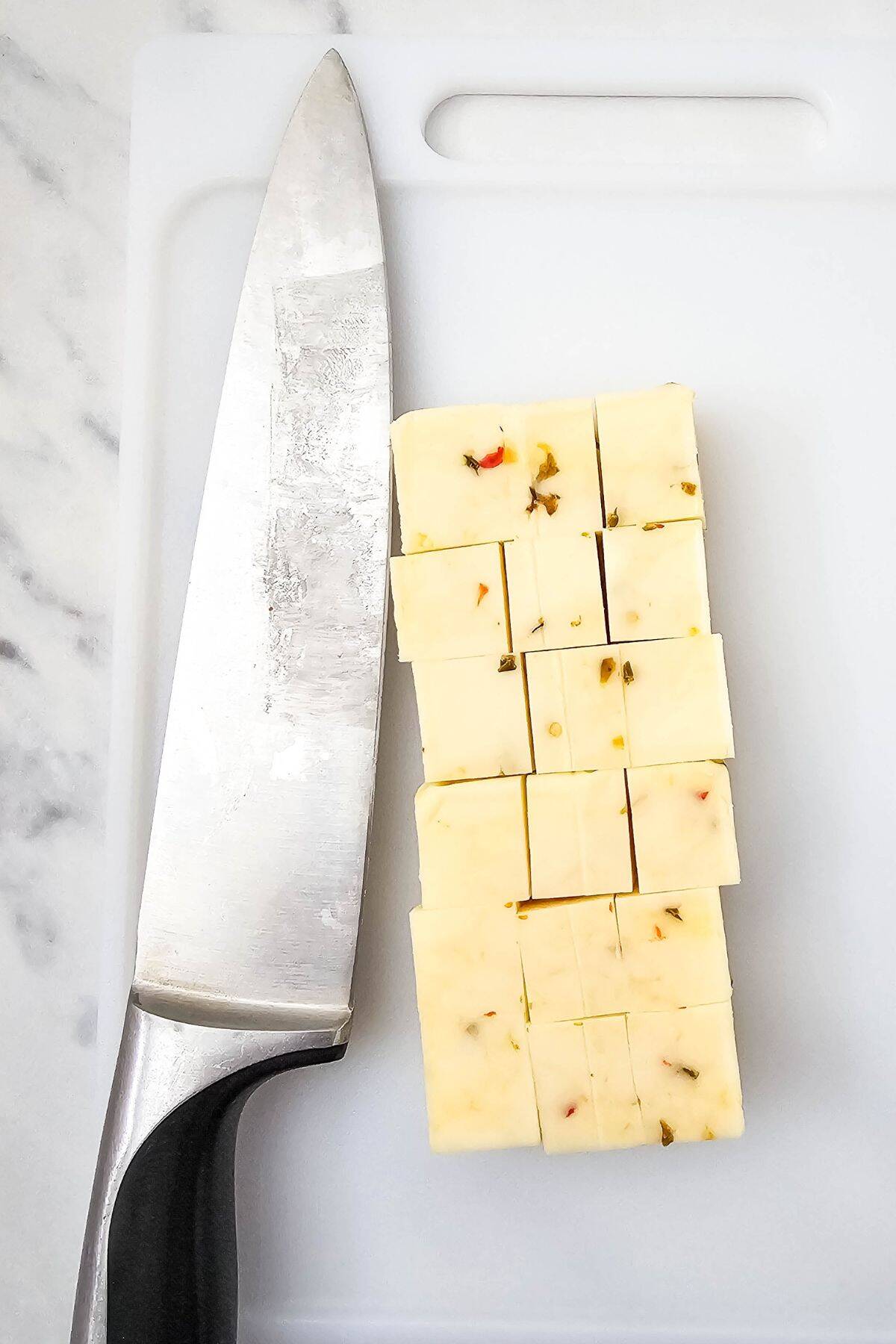 Sliced cheese cut into cubes, with a knife placed beside it on a cutting board.