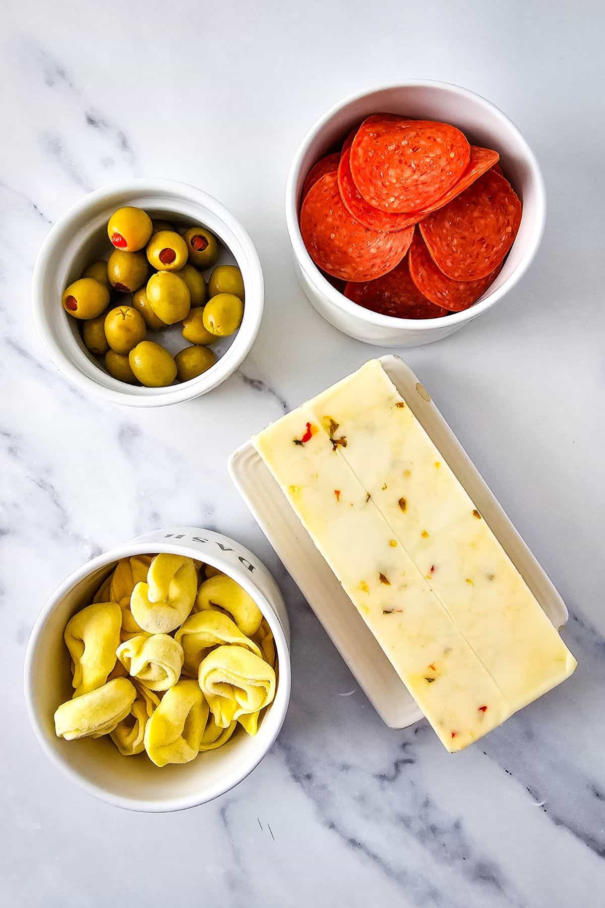 Ingredients for tortellini skewers—cooked tortellini, cubed cheese, sliced pepperoni, and olives—neatly arranged on a flat surface.