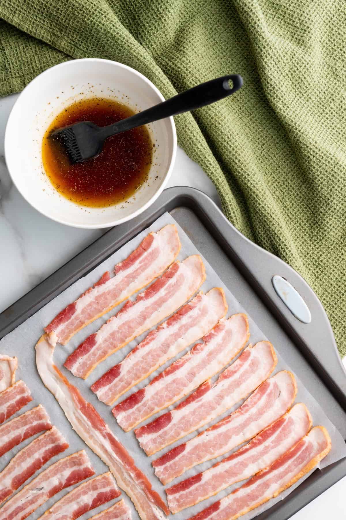 Maple brown sugar sauce in a small bowl with a brush, and bacon strips lined on a parchment-lined tray.