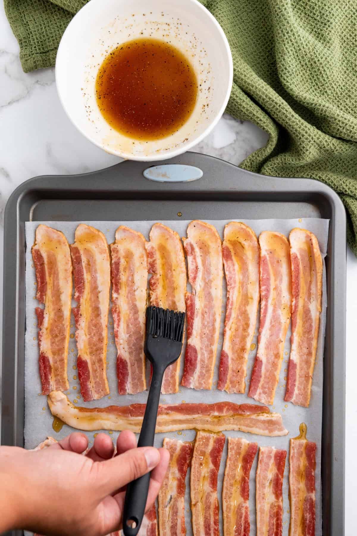 A hand brushing maple brown sugar sauce onto bacon strips on a parchment-lined tray.