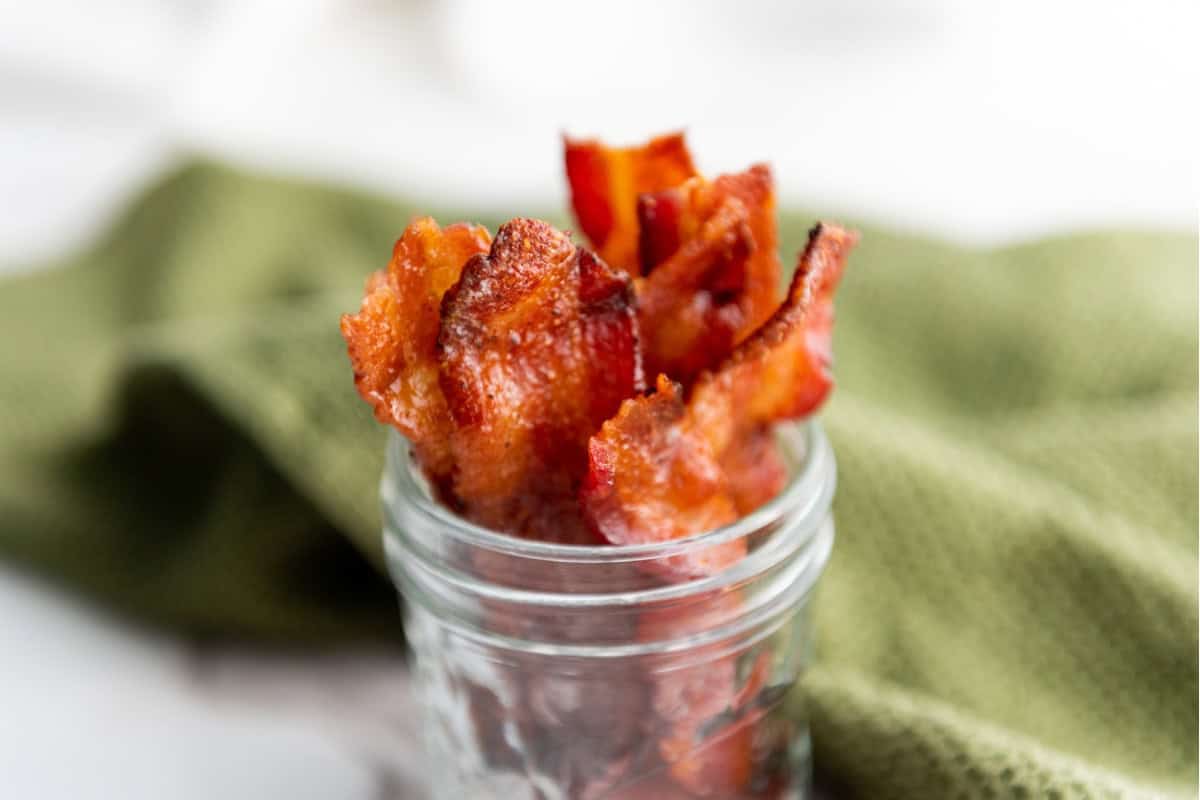 Candied maple brown sugar bacon in a small jar, with a green cloth in the background.