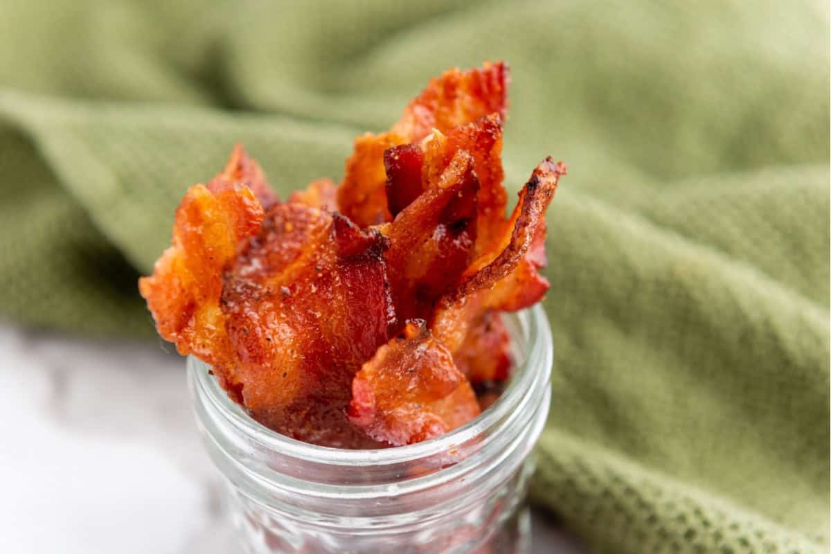 Candied maple brown sugar bacon in a small jar, with a green cloth in the background.