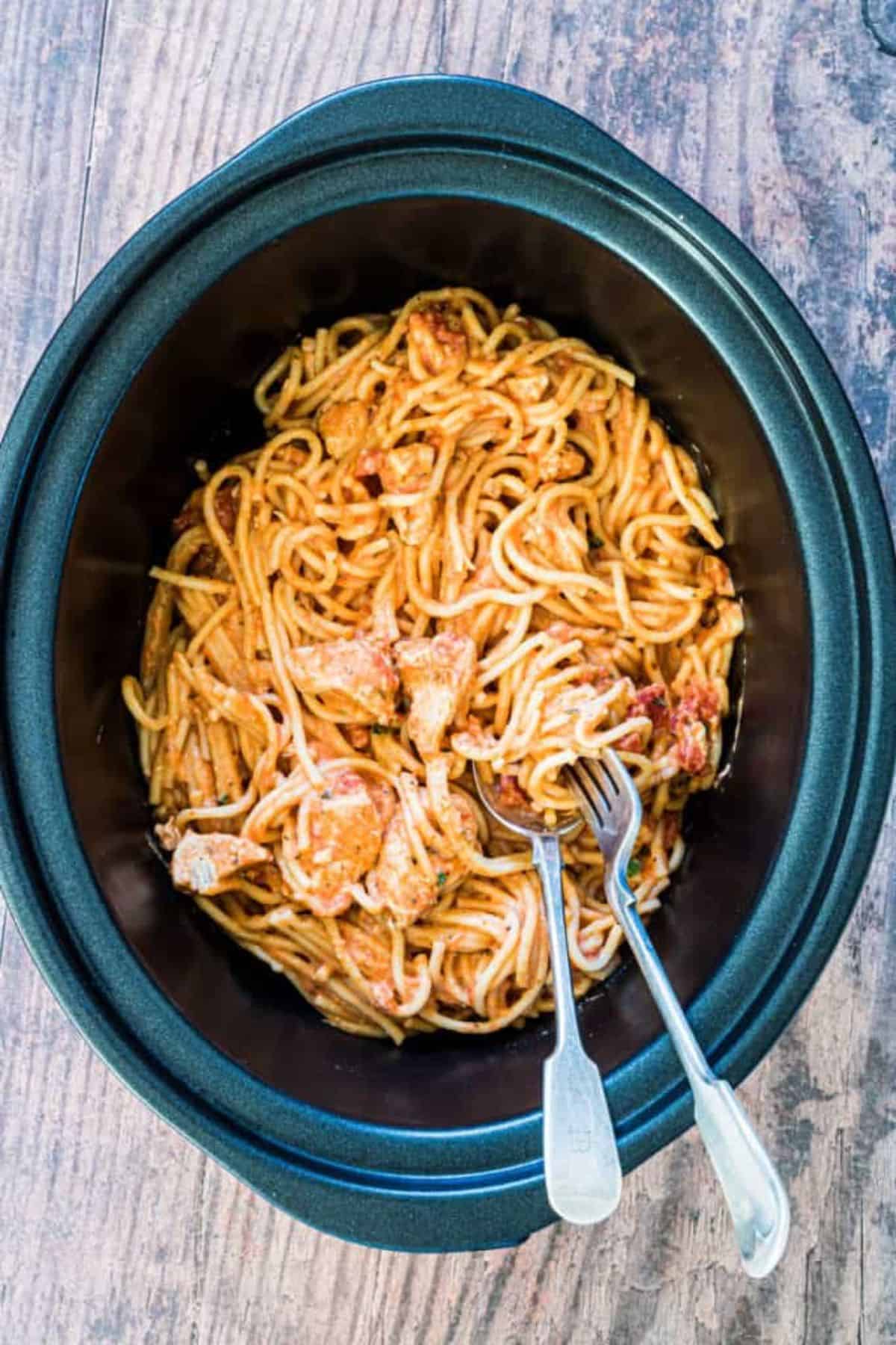 A crockpot with full of  slow cooker chicken spaghetti with spoon and fork resting on it.