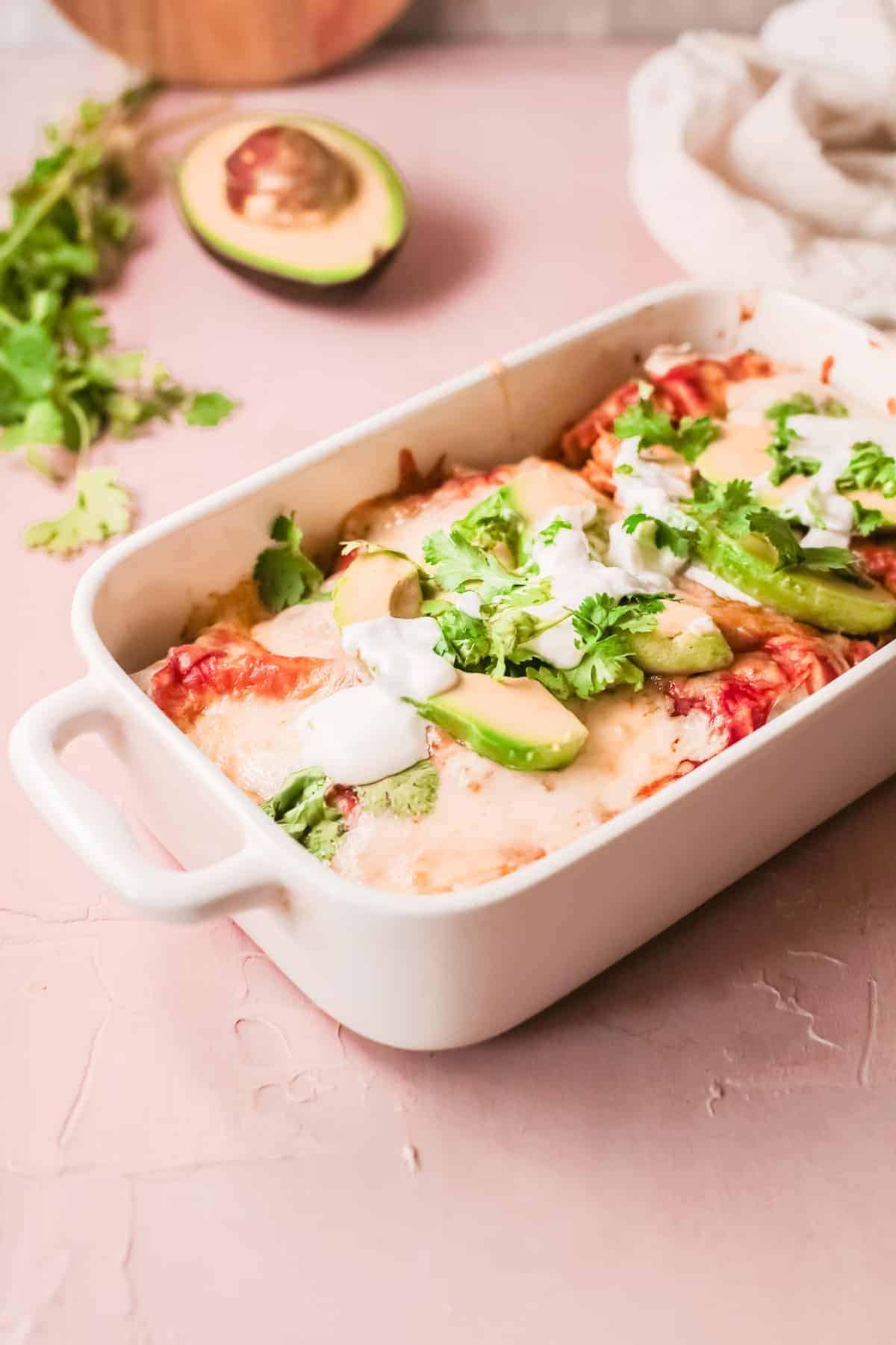 A serving dish with chicken enchiladas garnished with sliced avocado and parsley.