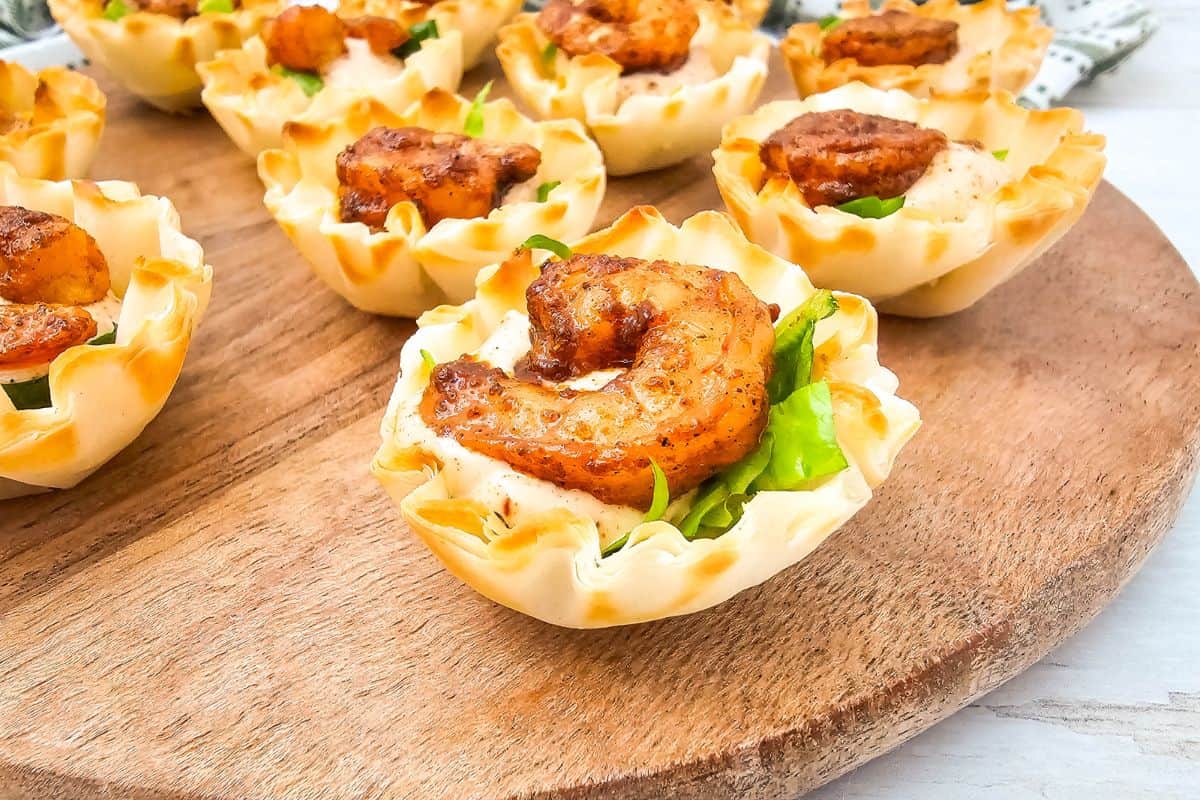 Close-up photo of chili lime shrimp cups arranged on a wooden board with shrimp, sauce, and lettuce in phyllo shells.