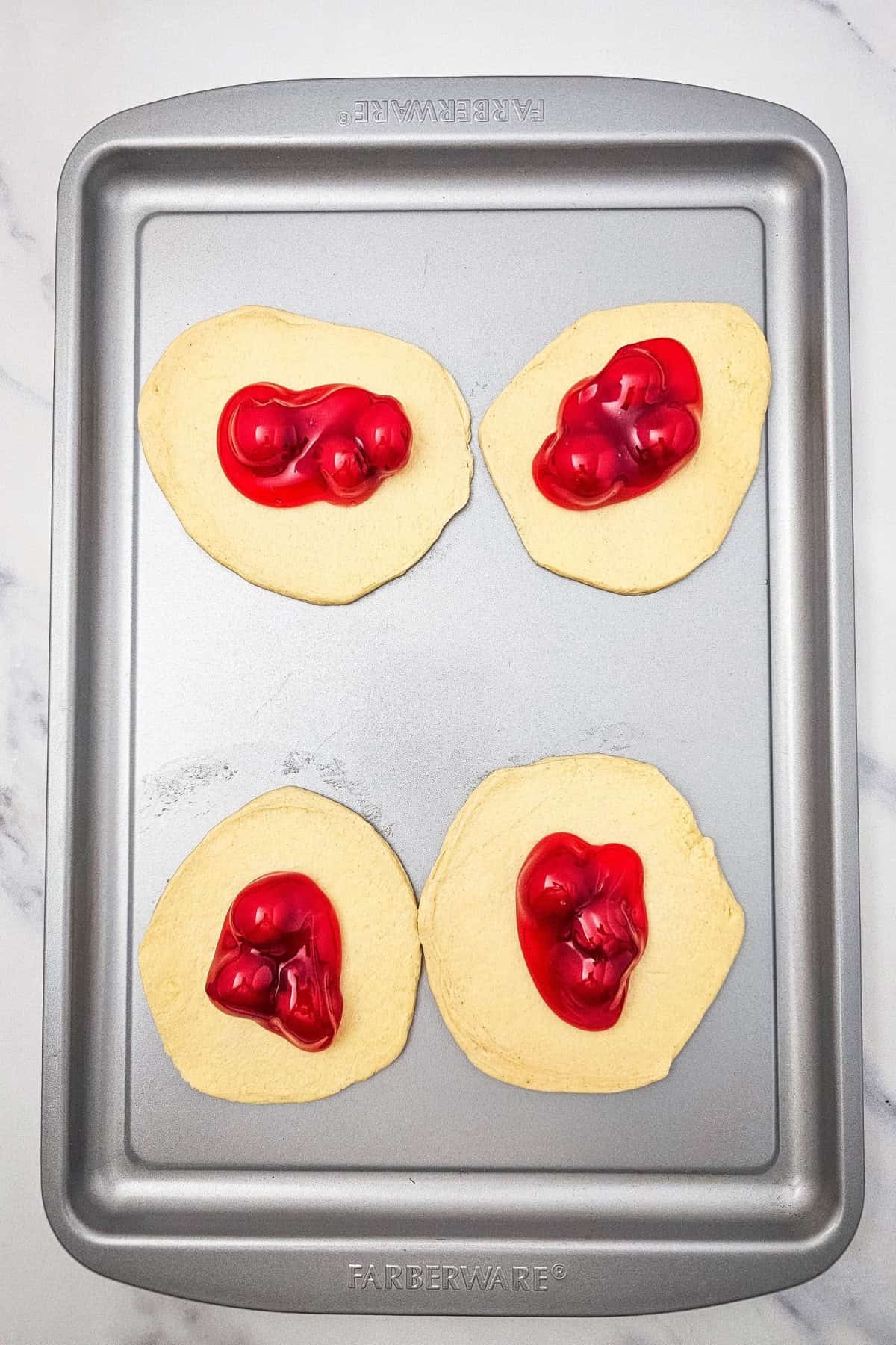 Flattened biscuit dough pieces lined up in the air fryer basket, each with cherry pie filling placed in the center.