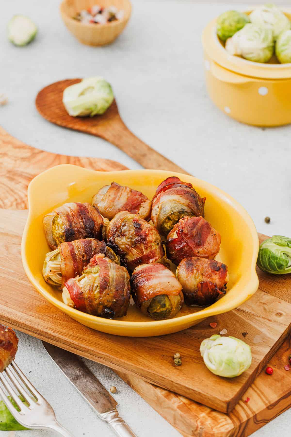 Maple-glazed bacon-wrapped Brussels sprouts served on a plate with a wooden board underneath, surrounded by fresh Brussels sprouts and herbs.