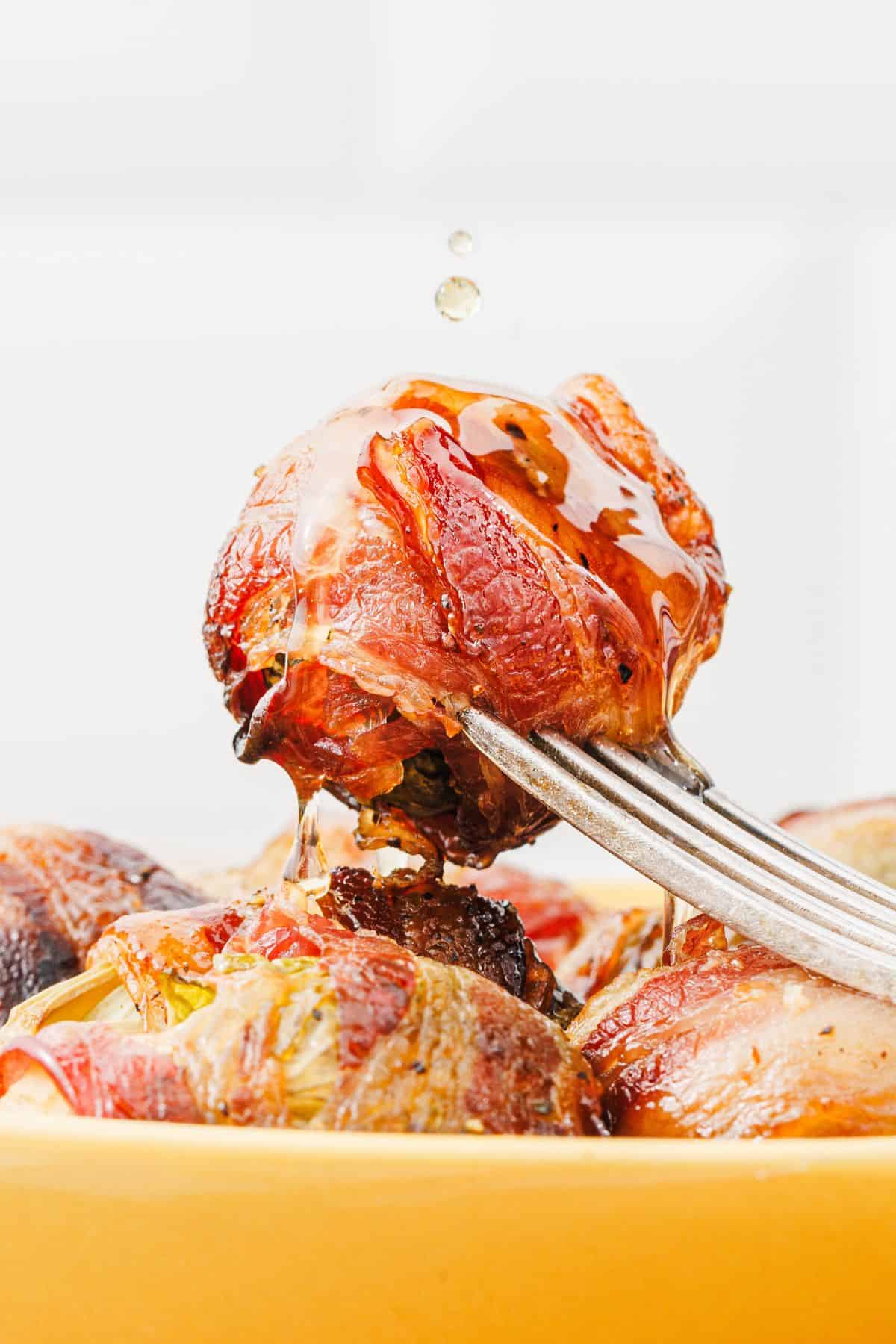 Close-up photo of a single maple-glazed bacon-wrapped Brussels sprout on a fork, with more wrapped sprouts arranged underneath on a serving plate.