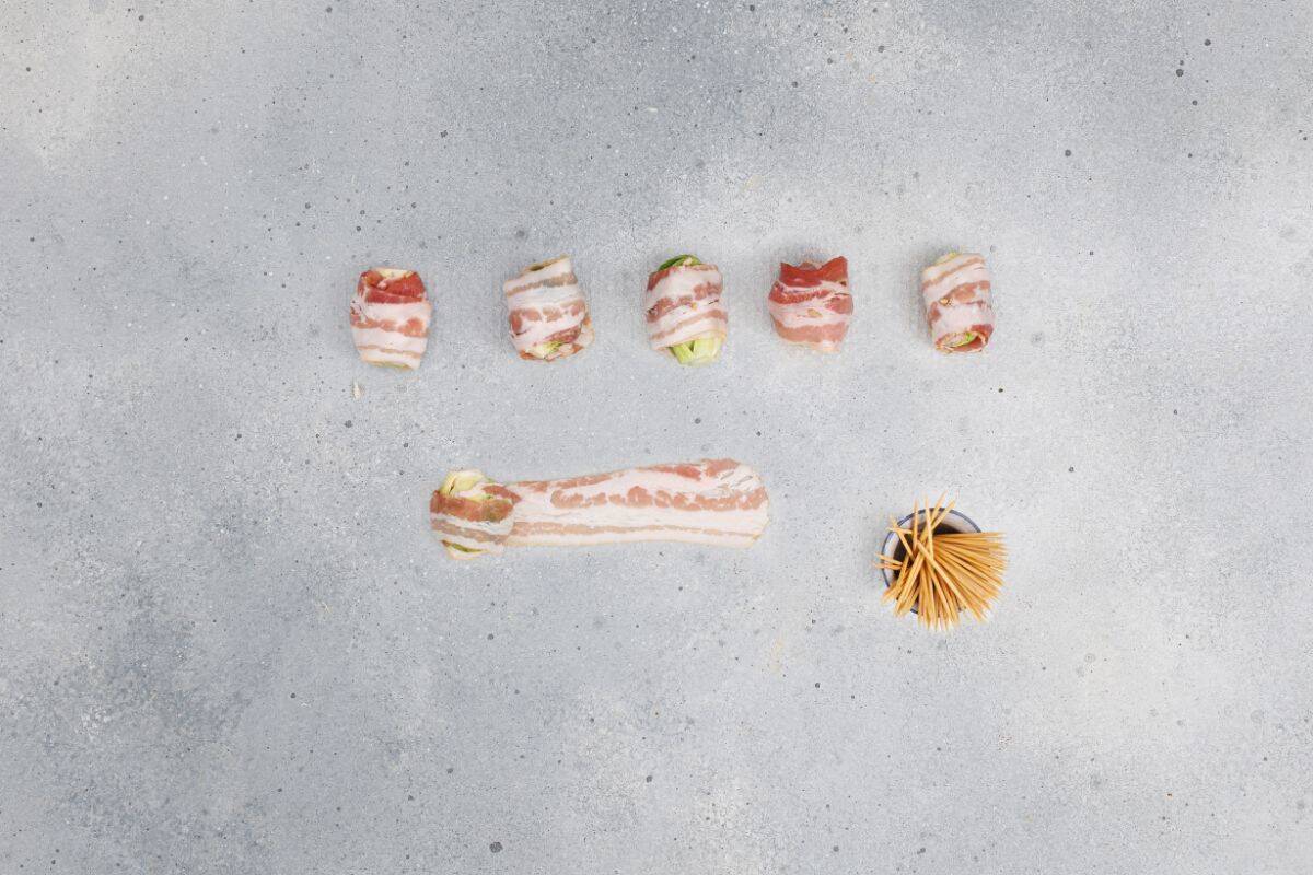 Bacon strips wrapped around Brussels sprouts, with other bacon strips laid flat on the surface nearby.