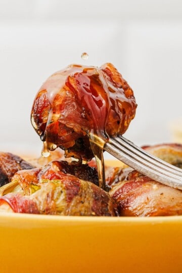 A single maple-glazed bacon-wrapped Brussels sprout on a fork, with more wrapped sprouts arranged underneath on a serving plate.