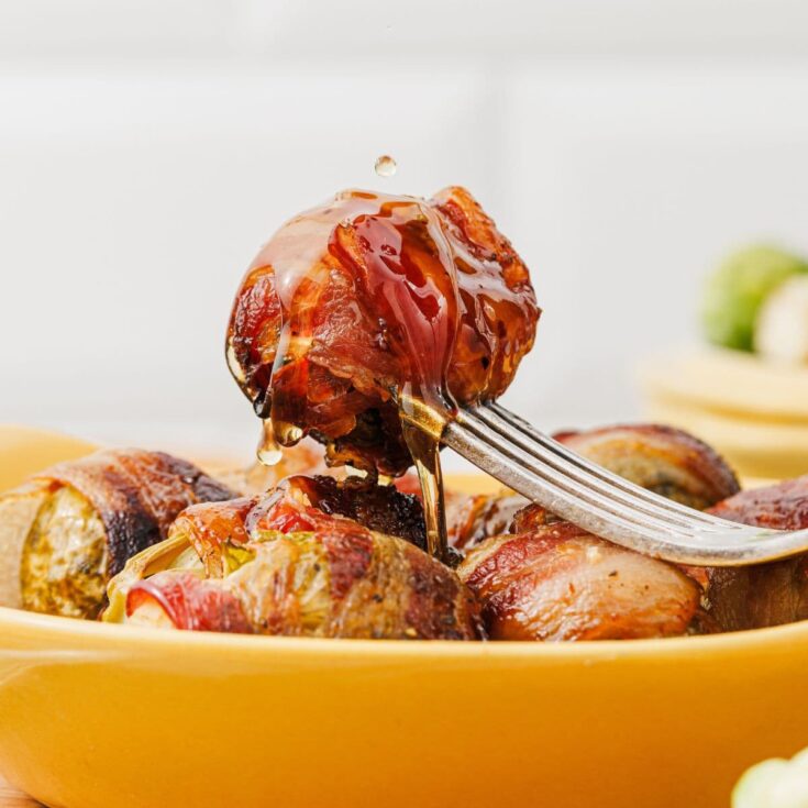 A single maple-glazed bacon-wrapped Brussels sprout on a fork, with more wrapped sprouts arranged underneath on a serving plate.