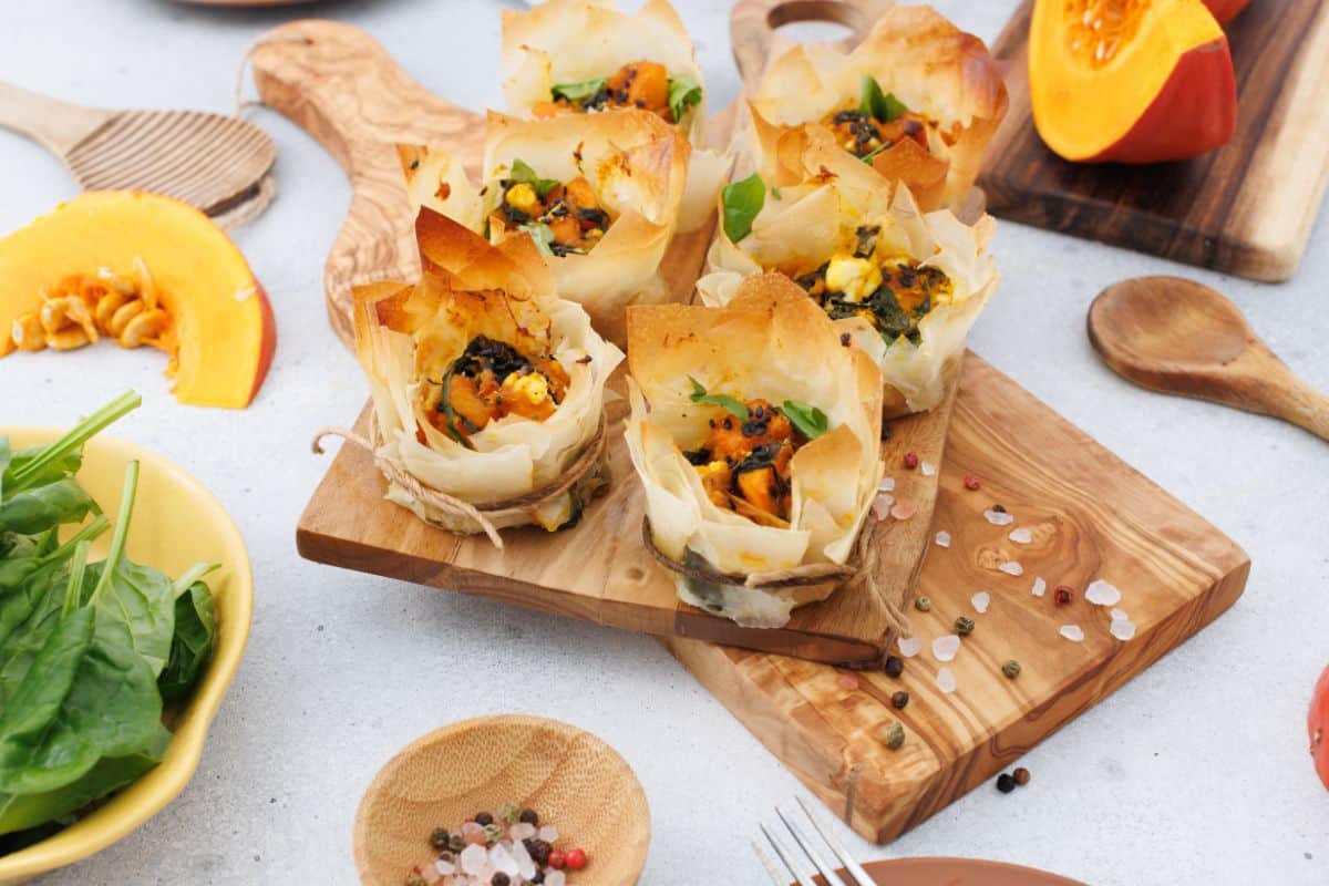 Pumpkin feta phyllo cups served on a wooden board, garnished with seeds, with a pumpkin and decorative items in the background.