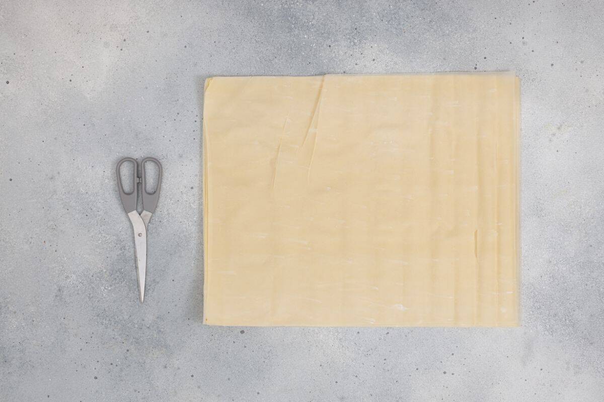 Thin sheets, likely phyllo dough, being cut into squares on a cutting board.