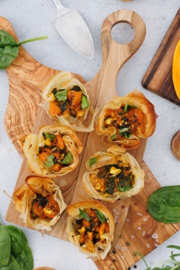 Pumpkin feta phyllo cups served on a wooden board, garnished with seeds, with a pumpkin and decorative items in the background.