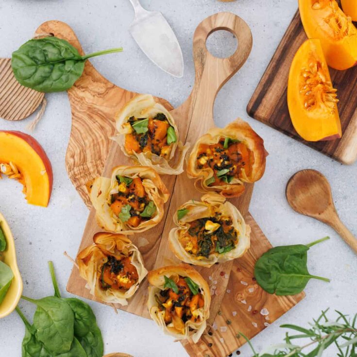 Pumpkin feta phyllo cups served on a wooden board, garnished with seeds, with a pumpkin and decorative items in the background.