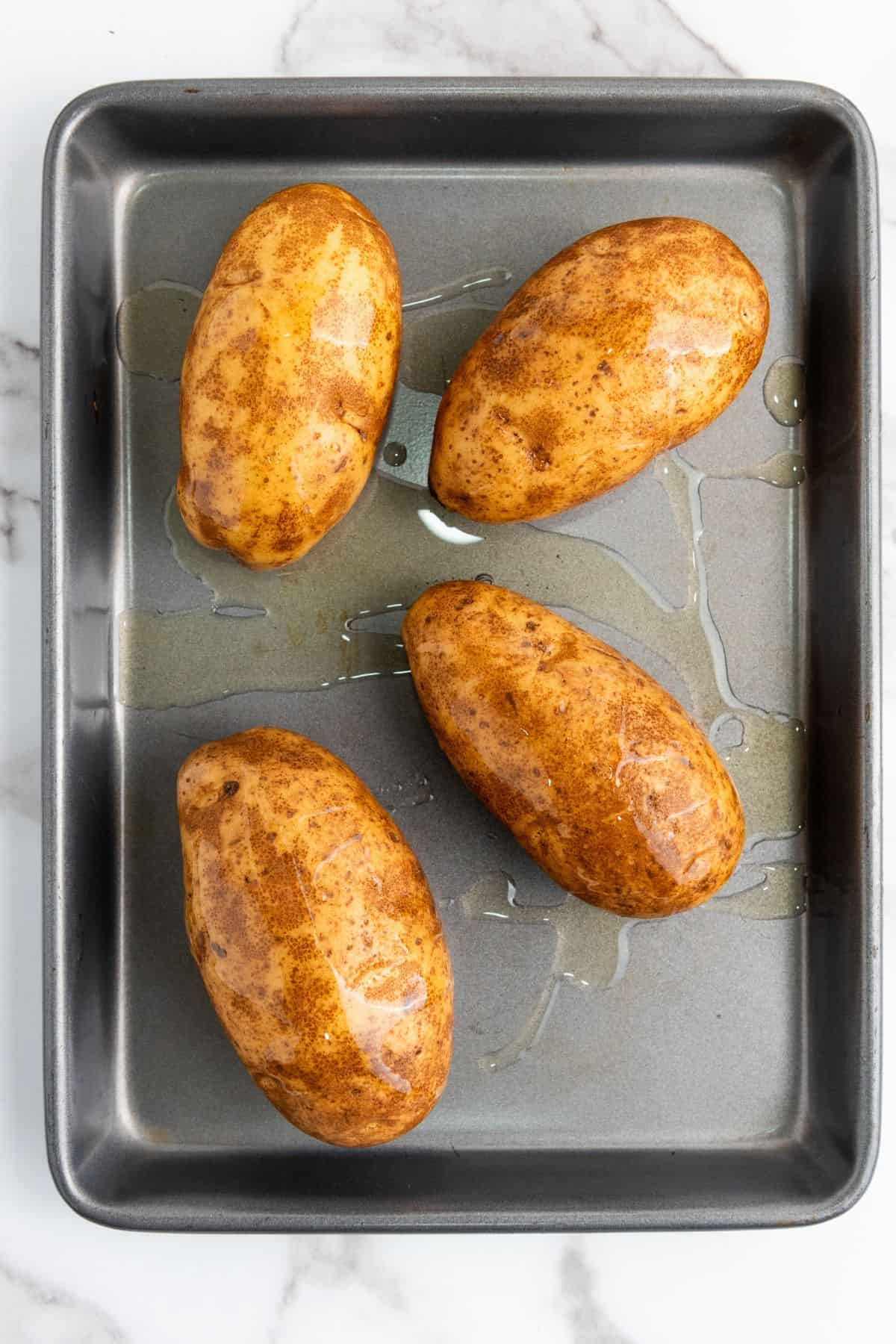 Four whole baked potatoes on a baking tray, uncut and with crispy skins.