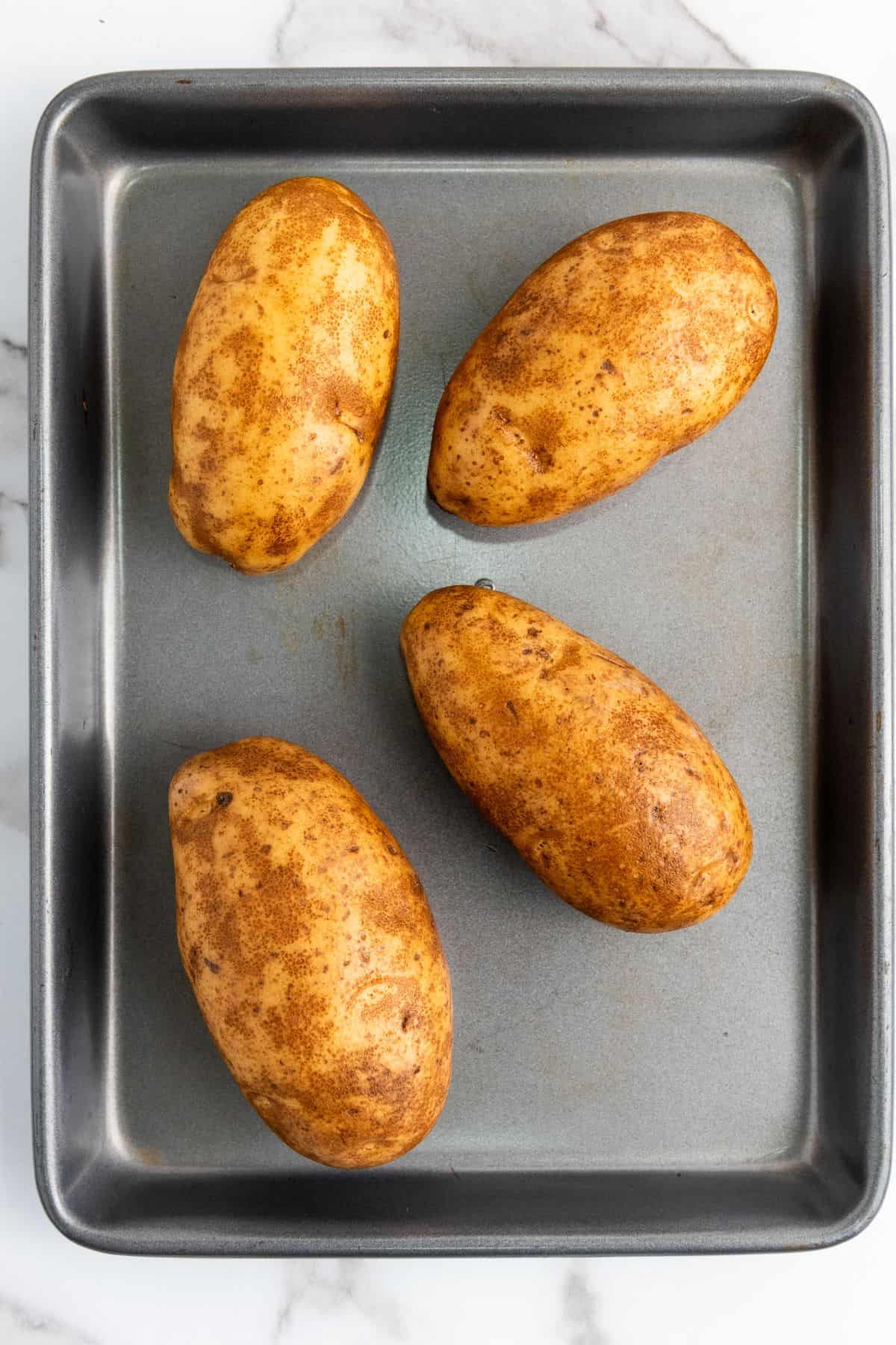 Four whole baked potatoes on a baking tray, uncut and with crispy skins.