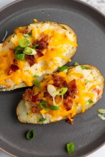 Two twice-baked potato halves served on a plate, topped with melted cheese, bacon bits, and sliced scallions.