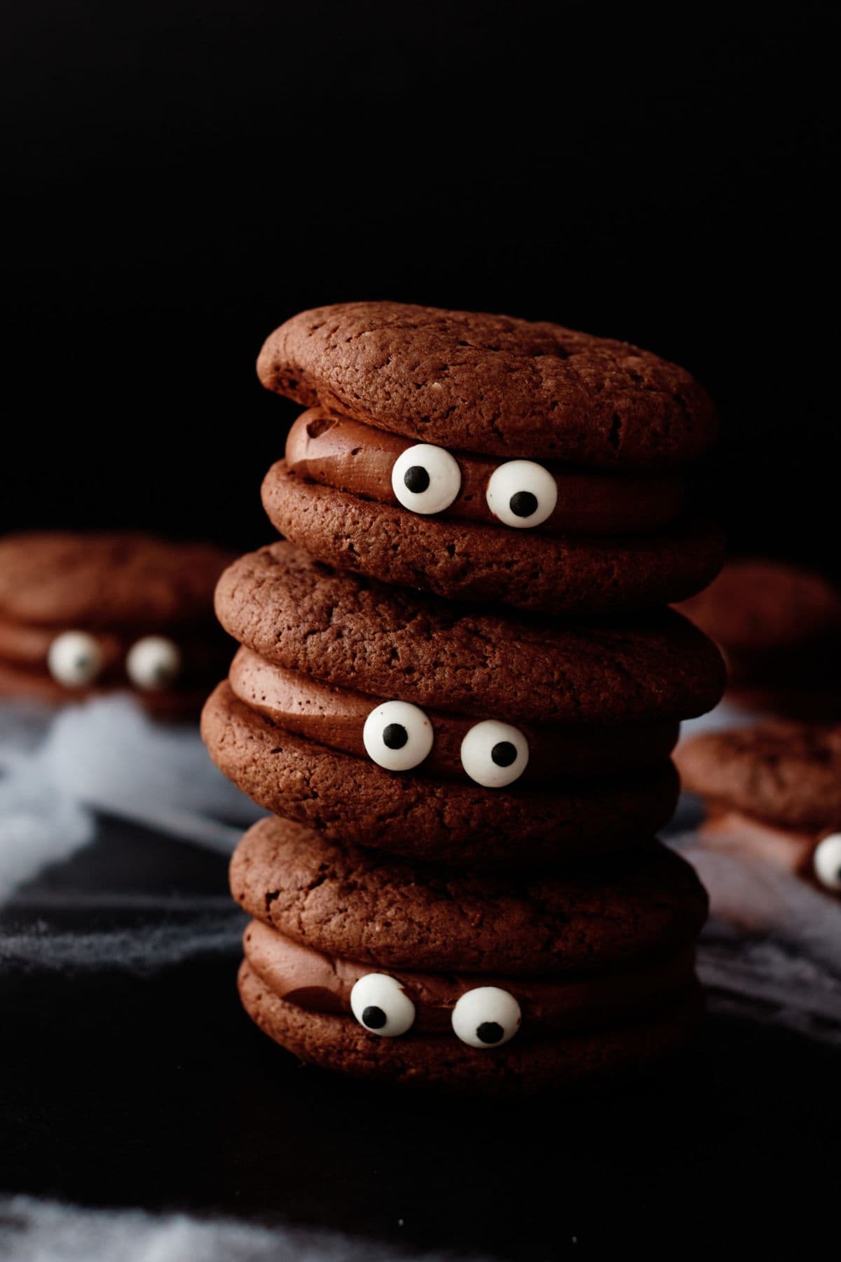 Stacked sandwich cookies decorated with candy eyes and chocolate coating, with more Halloween Eyeball Cookies scattered in the blurred background.