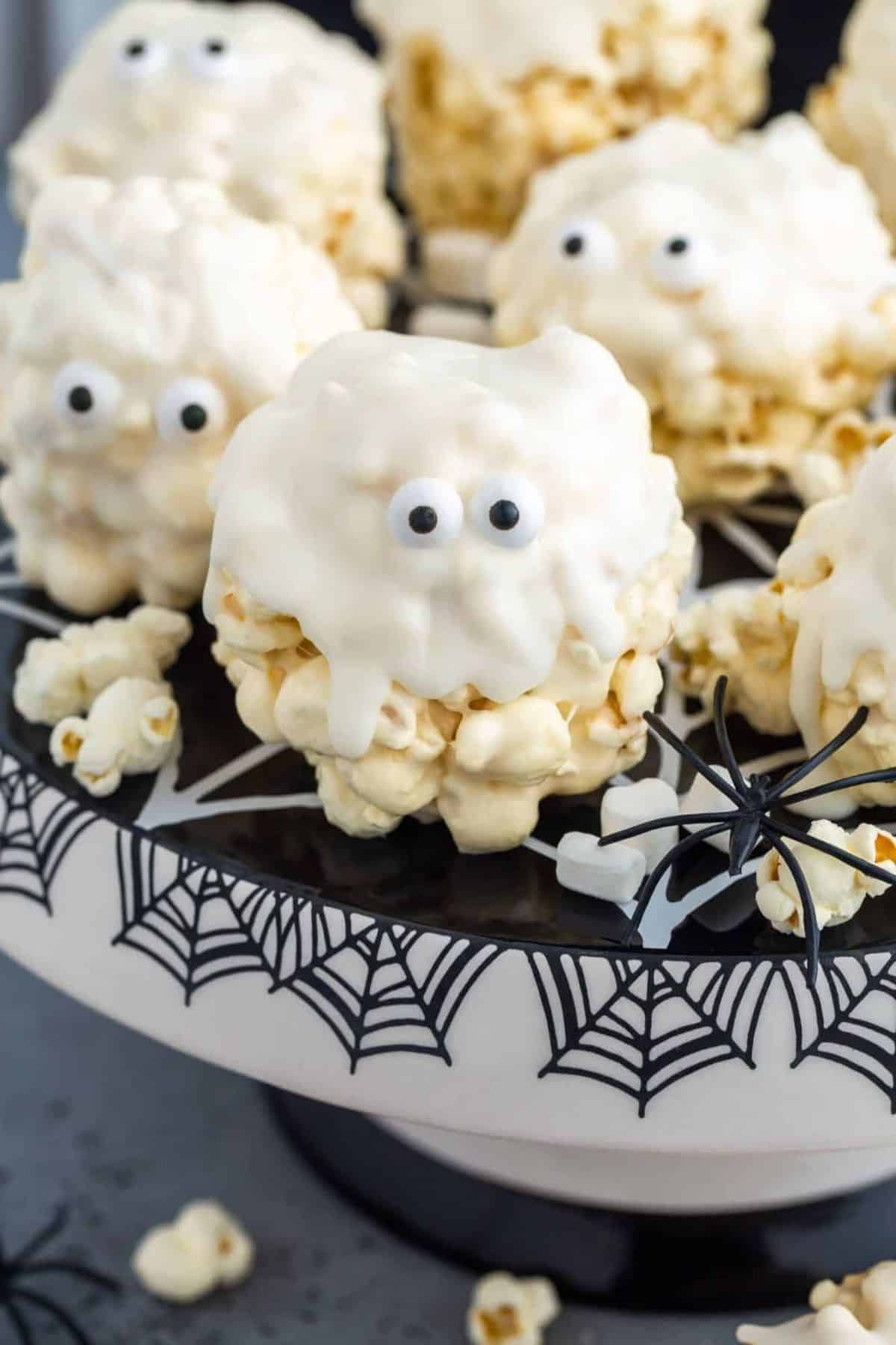 Close-up of marshmallow popcorn balls decorated like ghosts, served on a plate with candy eyes and a smooth white coating.