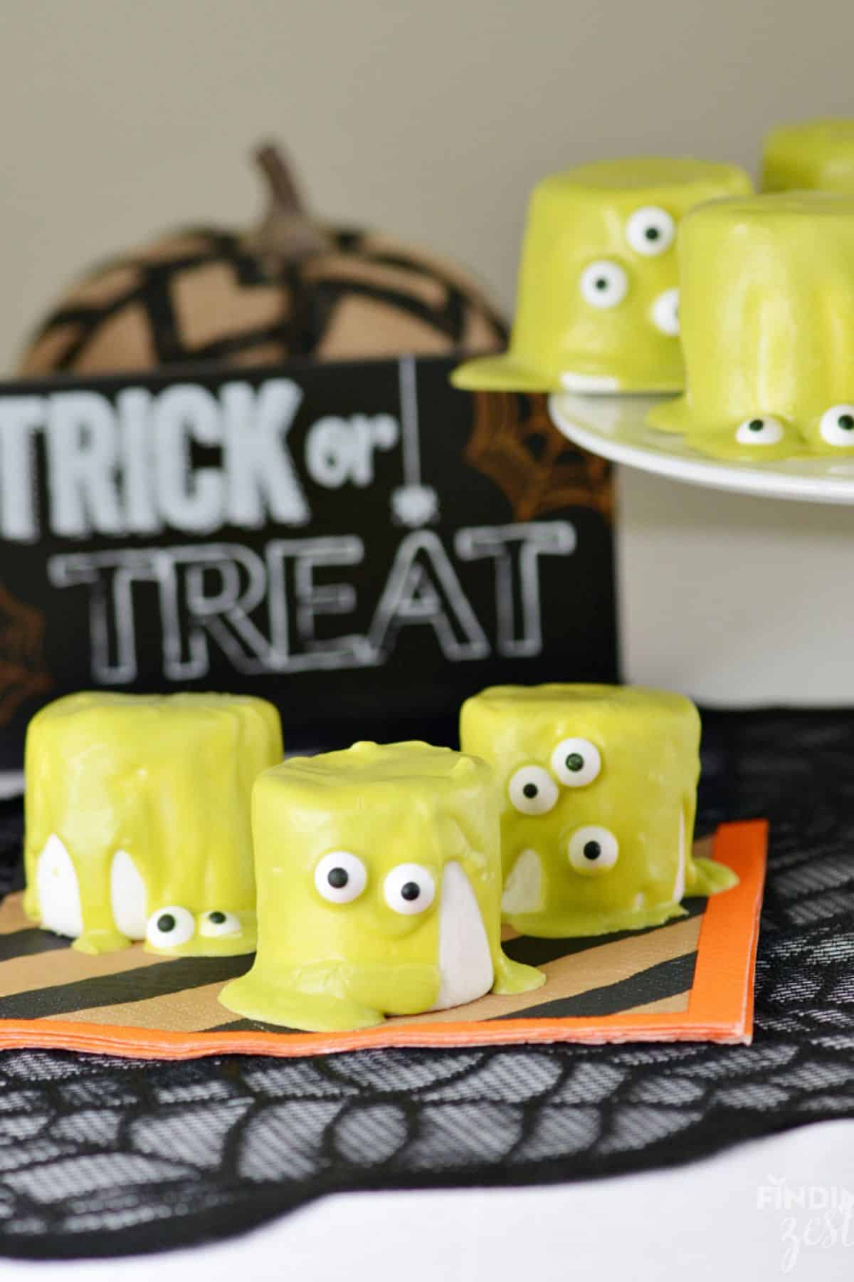 Green candy-coated marshmallows with candy eyes, arranged in front of a Halloween banner hanging in the background.