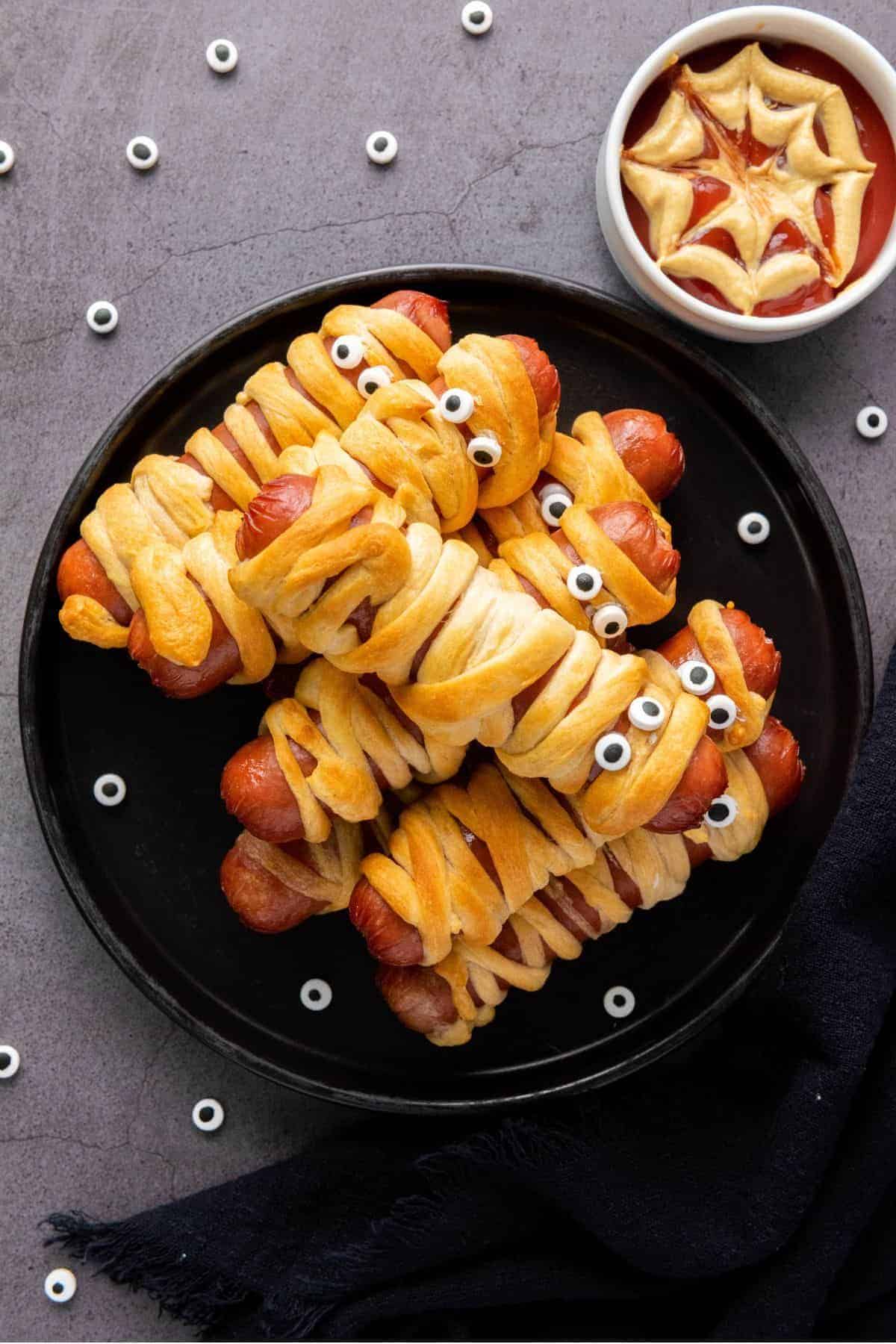Golden crescent-wrapped mummy dogs arranged on a plate with a spooky spider web dip on the side.