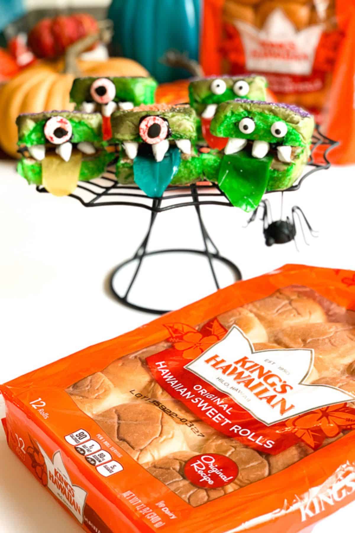 Assorted monster-themed treats displayed on a spider web-shaped stand with packed dinner rolls placed nearby on the table.