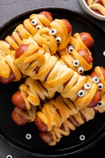 Golden brown mummy hotdogs wrapped in crescent roll strips with edible eyes, arranged on a serving plate with dipping sauce on the side.