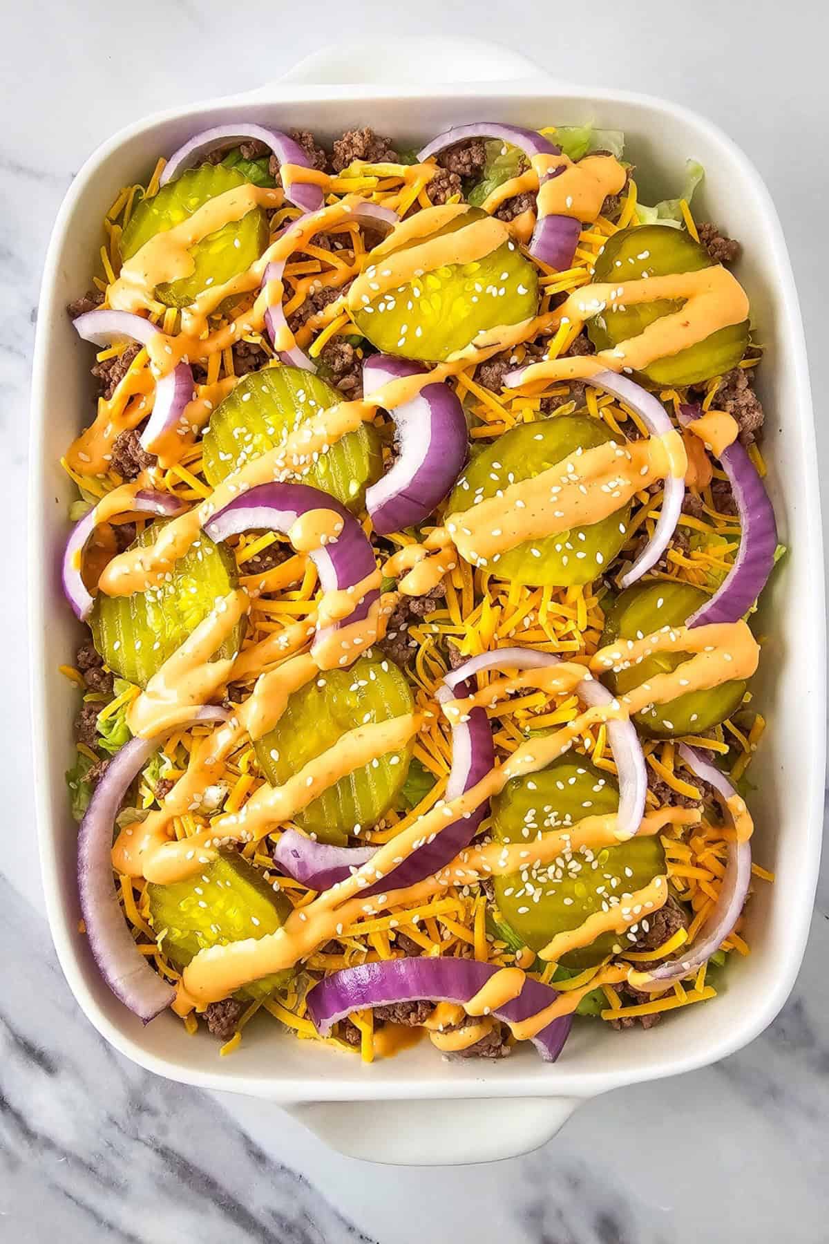 A serving dish with lettuce, cheese, ground beef topped with onion and pickles, drizzled with dressing.