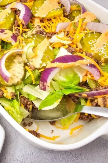 A serving dish with Big Mac Salad, topped with beef, lettuce, pickles, cheese, and dressing, with a spoon on the side.