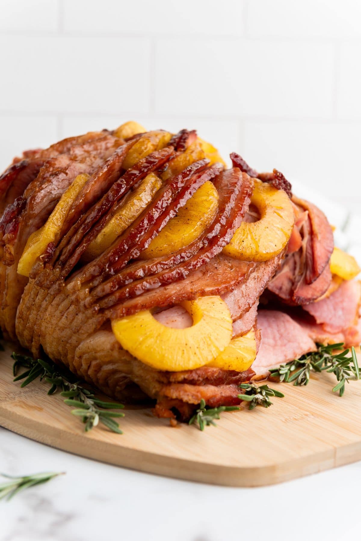 A serving of brown sugar pineapple ham sliced and arranged, garnished with pineapple rings, a light glaze, and sprigs of rosemary around the edges.