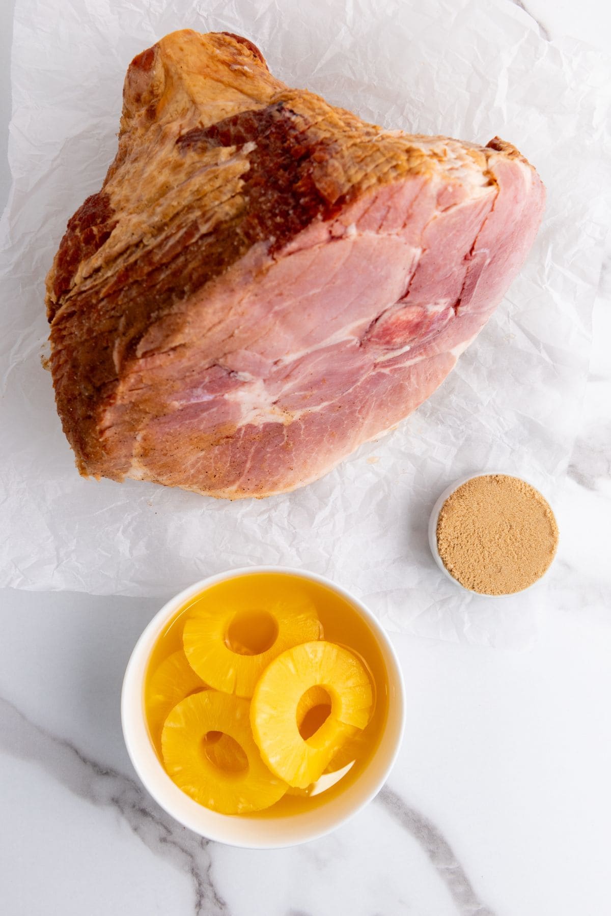 A can of pineapple slices, a spiral ham, and a small bowl of brown sugar arranged on a kitchen counter.