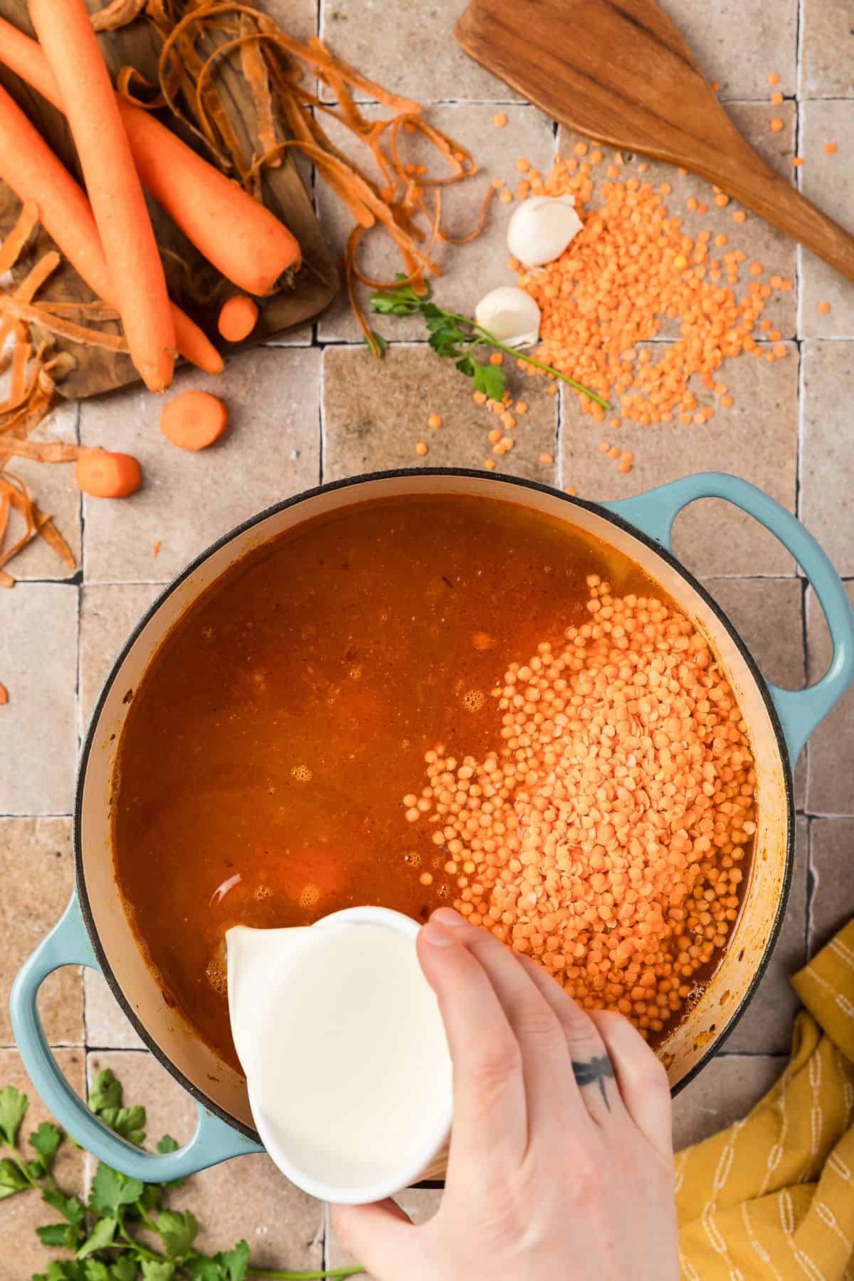 A hand pouring broth and milk into a saucepan with carrots and onions.