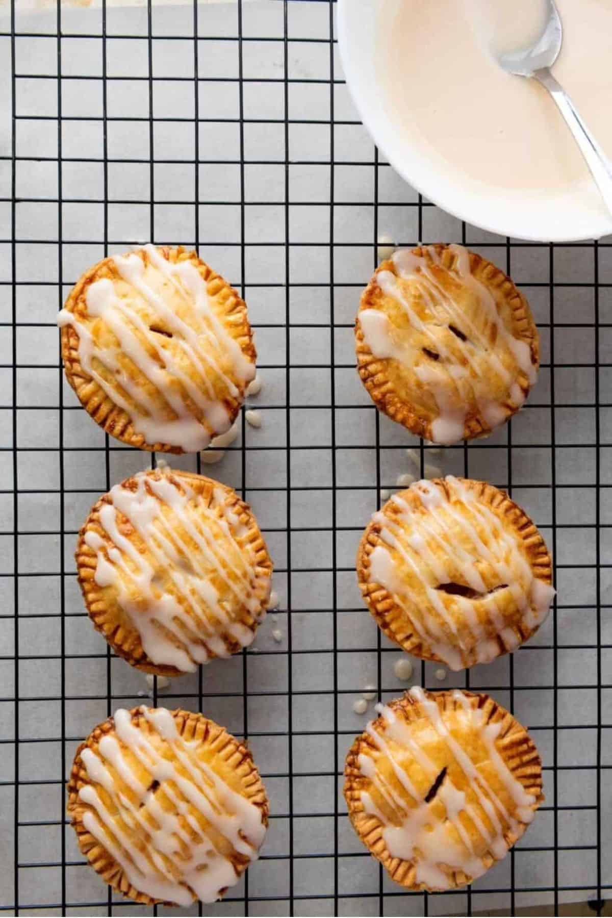 Apple hand pies lined up on a cooling rack, with a bowl of melted glaze nearby.