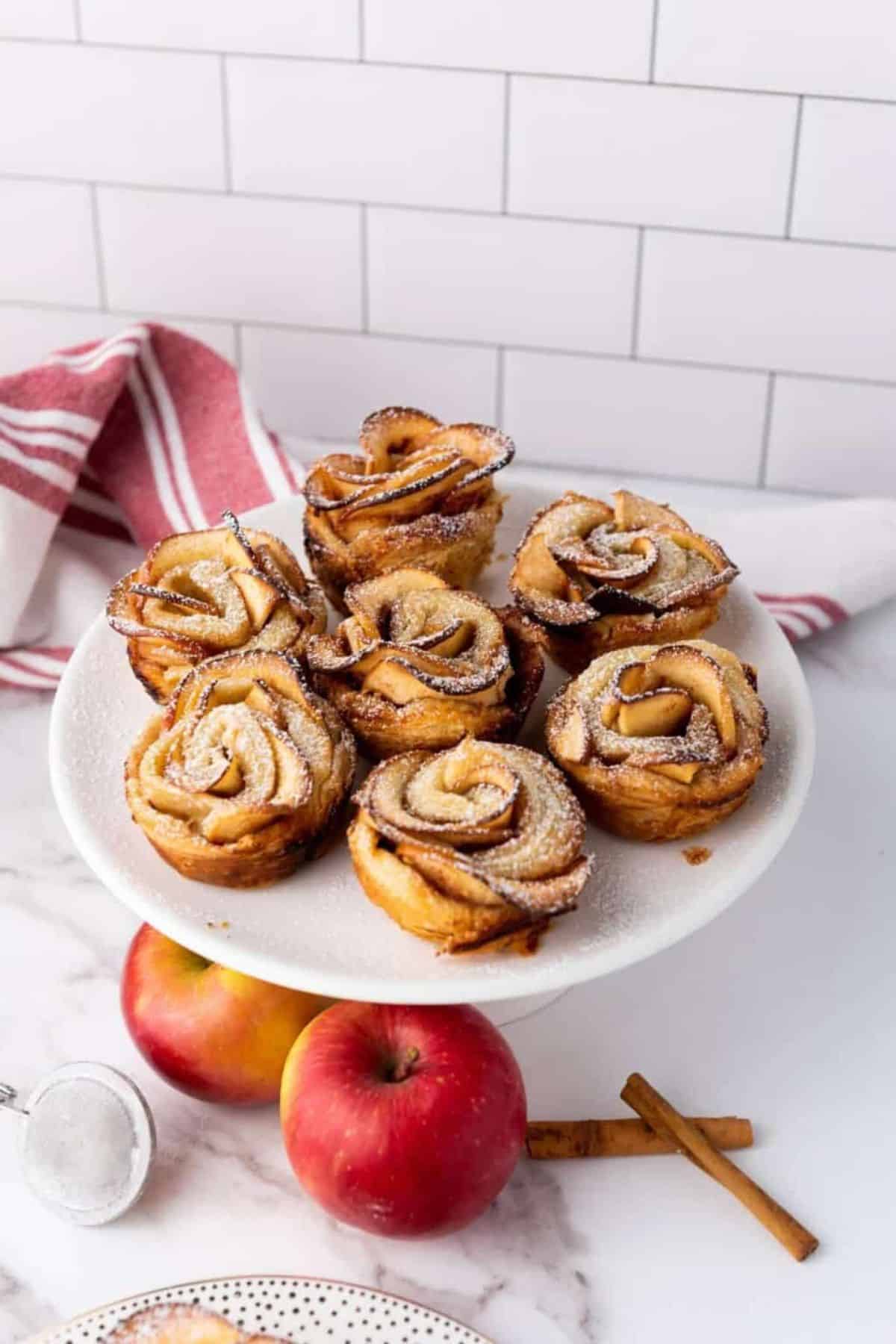 Puff pastry apple roses arranged on a cake stand with fresh apples placed below.