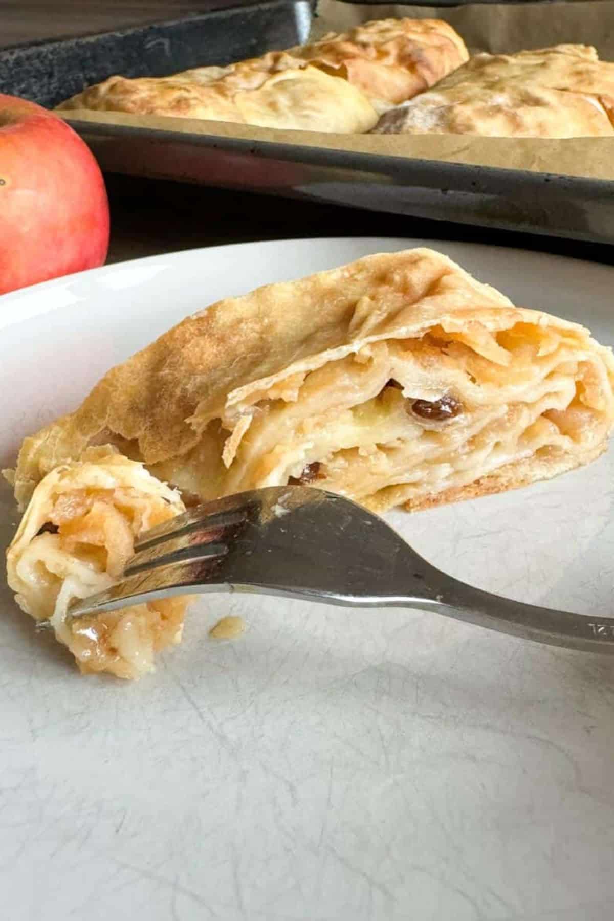 Slice of apple strudel served on a plate, with a fork pinching a bite from the pastry.