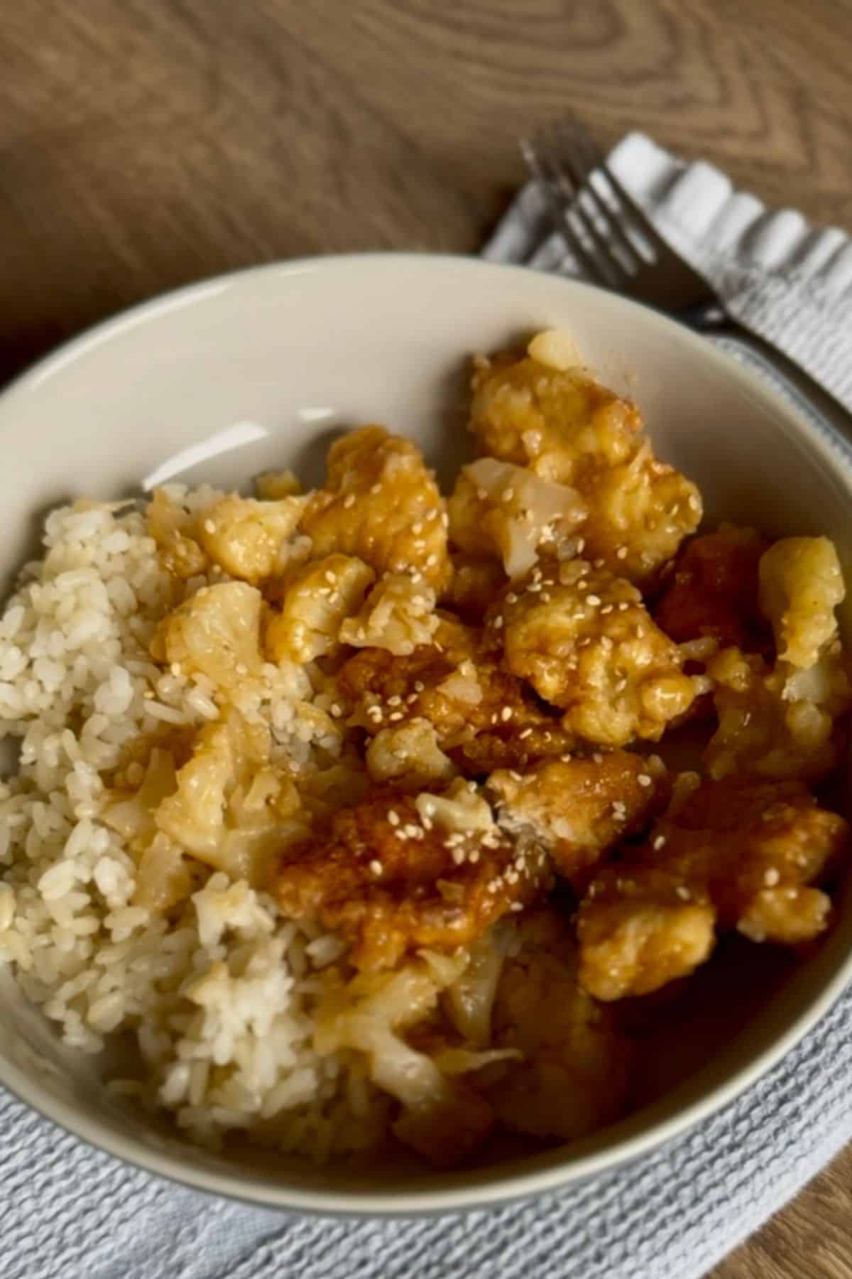 A plate with crispy fried sweet and sour chicken with cauliflower over rice resting on a cloth.