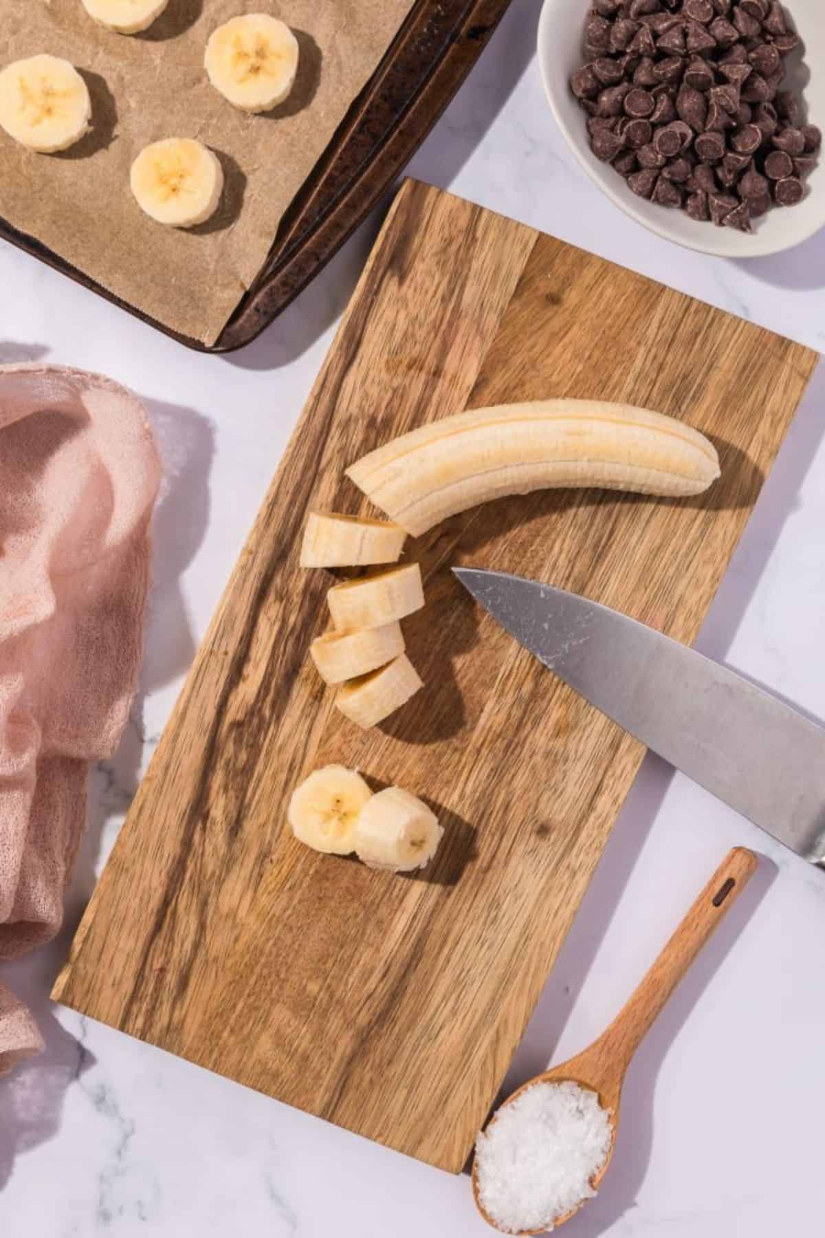 A banana on a chopping board being sliced into pieces with a knife placed beside it.