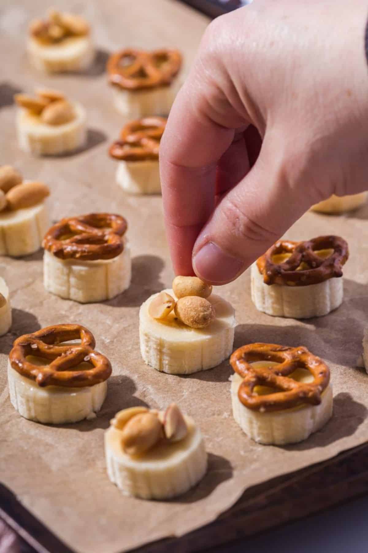 A hand placing cocktail peanuts on top of sliced banana pieces arranged on parchment paper.