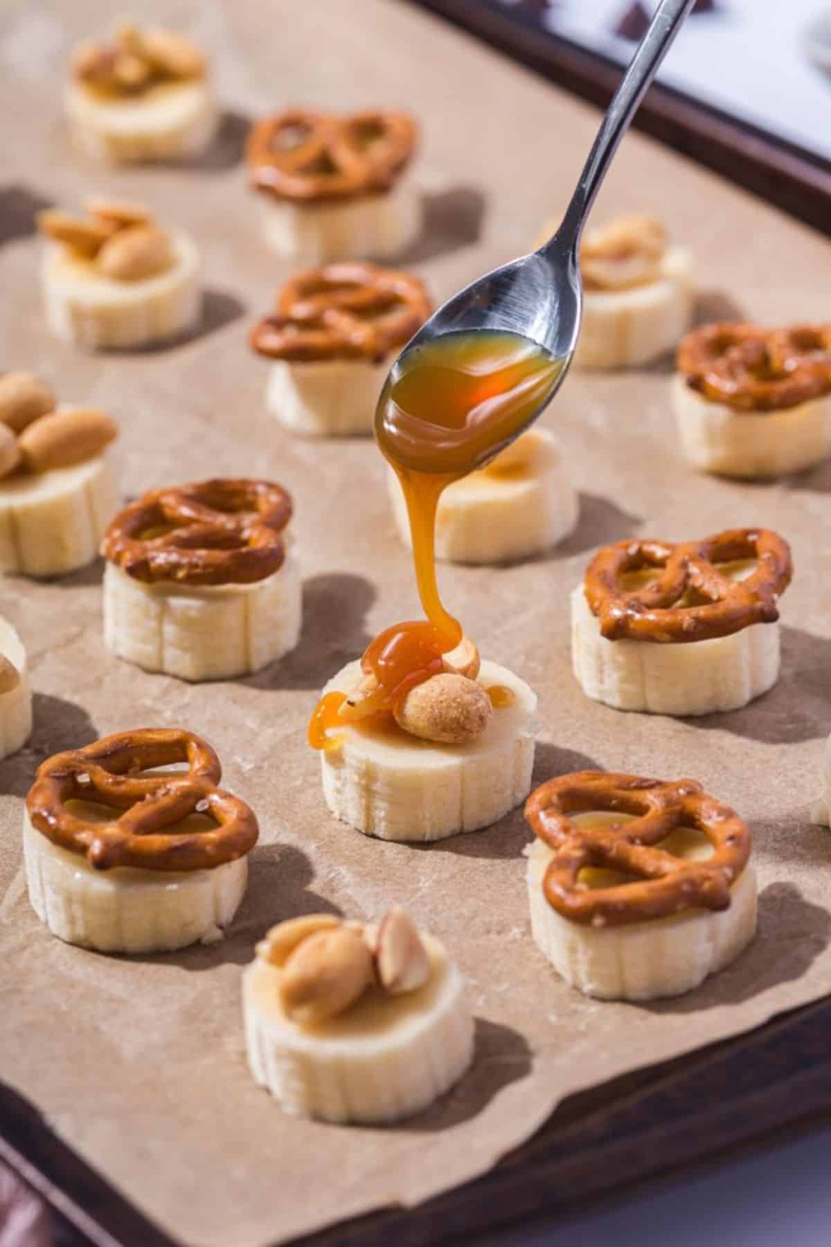 A spoon drizzling caramel sauce over sliced banana pieces arranged on parchment paper.